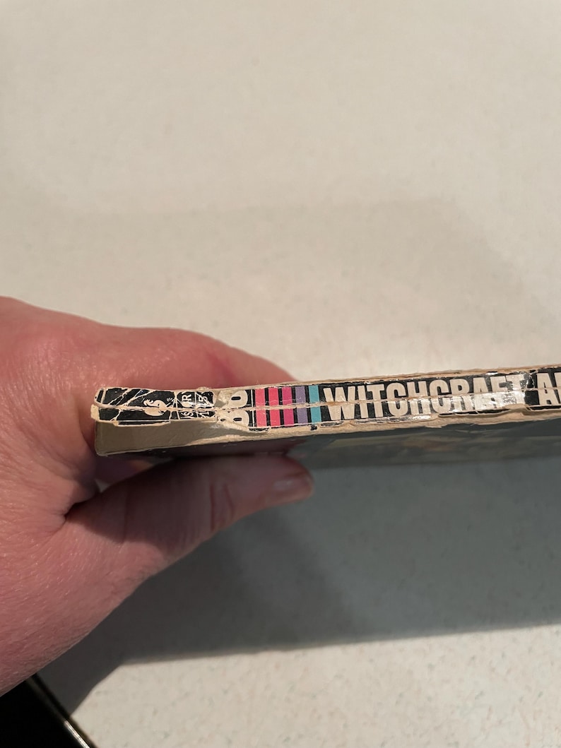 K&ouml;nnte beinhalten: Buchr&uuml;cken mit dem Titel "WITCHCRAFT" in wei&szlig;en Buchstaben. Der R&uuml;cken hat eine Reihe vertikaler Streifen in Rot, Rosa, Blau und Lila. Die Kanten des Buches weisen Gebrauchsspuren auf.