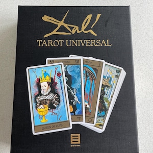 Puede incluir: Una caja negra de cartas del tarot con letras doradas que dicen "Dali Tarot Universal". Varias cartas del tarot están abanicadas, mostrando ilustraciones coloridas. Una carta muestra a una reina sosteniendo una copa dorada. La caja tiene una apariencia texturizada.