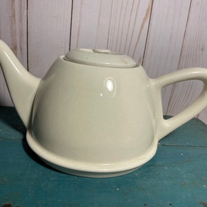 Peut inclure: Une théière en céramique blanche avec un corps arrondi et un bec légèrement incurvé. La théière a une finition lisse et mate et un design simple.