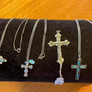 Könnte beinhalten: Fünf silberne Halsketten mit verschiedenen Kreuzanhängern. Die Kreuze sind mit Türkis und klaren Strasssteinen verziert. Ein Kreuz zeigt eine Jesusfigur.