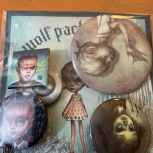 May include: A collection of art buttons and a small square print featuring surreal illustrations. The buttons showcase portraits of stylized figures with dark features and a doll-like appearance. The background art has a whimsical, fantasy theme.