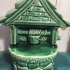 Peut inclure: Puits de souhaits en céramique verte avec un seau et une corde. Le puits a un toit et une base en pierre. Le texte "Wishing you well" est inscrit sur la base.