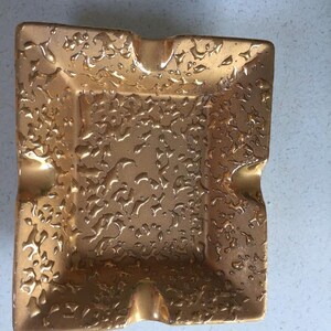 May include: A gold-colored ceramic ashtray with a decorative embossed floral pattern.