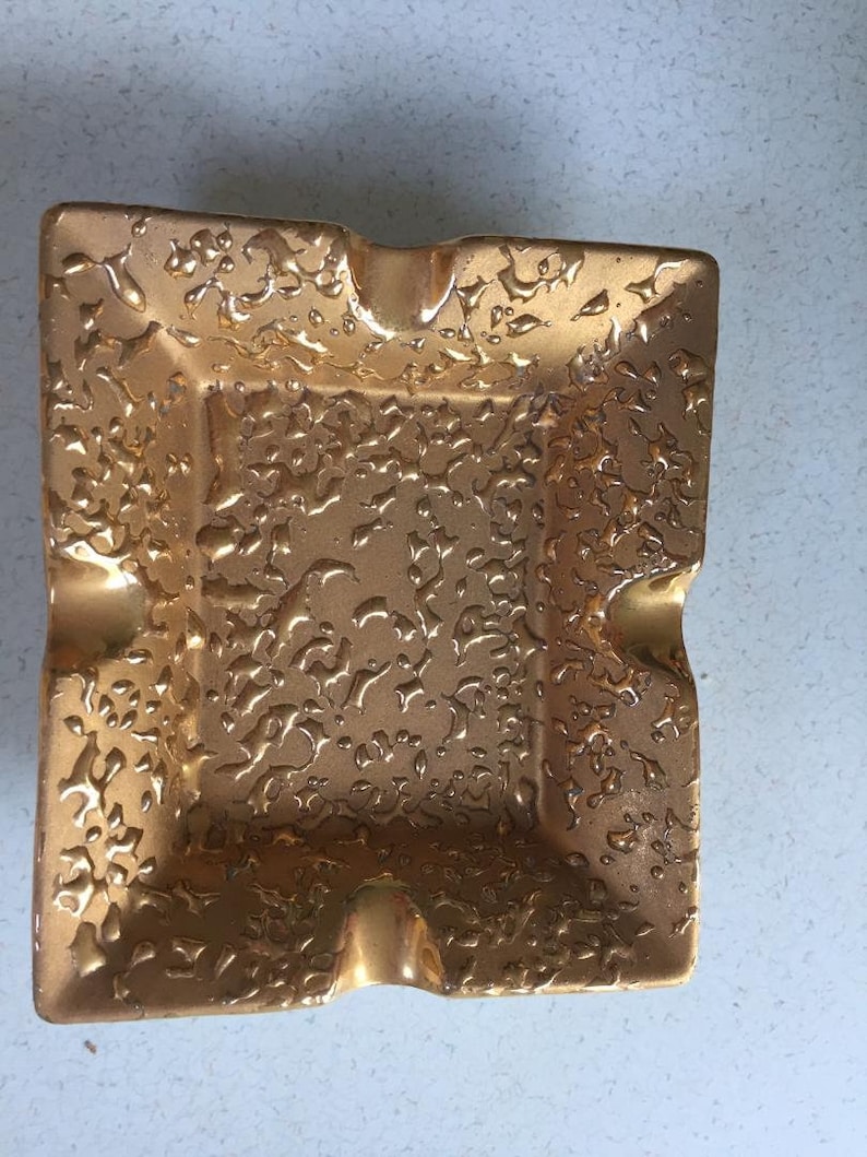 May include: A gold-colored ceramic ashtray with a decorative embossed floral pattern.