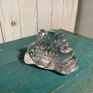 May include: A clear glass sculpture of a frog with a textured surface. The frog is sitting on a blue wooden surface.