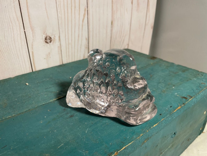 May include: A clear glass sculpture of a frog with a textured surface. The frog is sitting on a blue wooden surface.