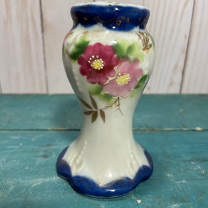 May include: A small white ceramic vase with a blue and gold rim and floral design. The vase features pink flowers with yellow centres and green leaves.