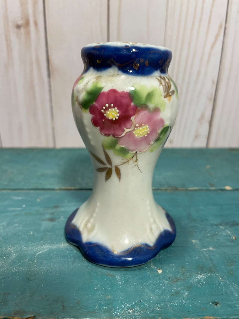 May include: A small white ceramic vase with a blue and gold rim and floral design. The vase features pink flowers with yellow centres and green leaves.