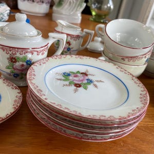 Puede incluir: Un juego de té de porcelana vintage con diseños florales. El juego incluye una tetera, una jarra de crema, un azucarero, tazas de té, platillos y un plato. Las tazas de té y los platillos tienen un delicado patrón floral con acentos rosados, azules y rojos.