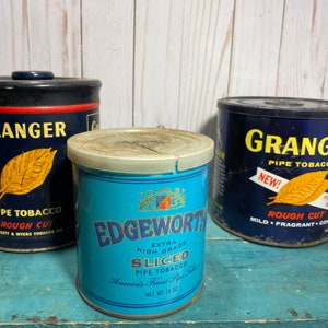 Puede incluir: Tres latas de tabaco vintage. Dos son azules con letras doradas y dicen "Granger Pipe Tobacco" y "¡Nuevo! Corte áspero Suave Fragante Fresco". La tercera lata es azul y blanca y dice "Edgeworth Extra Alta Calidad Rebanadas Tabacco para pipa El mejor tabaco para pipa de América Peso neto 14 oz."