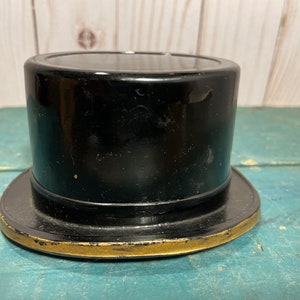 Puede incluir: Un sombrero de copa negro con ala ancha y ribete dorado. El sombrero es cilíndrico con la parte superior plana y un ala ligeramente curvada. El sombrero parece estar hecho de un material brillante.