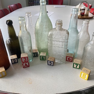 Puede incluir: Una colección de botellas de vidrio vintage de diversas formas y tamaños, dispuestas sobre una mesa blanca con bloques de madera numerados del 1 al 9. Las botellas son principalmente de vidrio transparente con algunos matices verdes y marrones. Los bloques están dispuestos en línea, con los números mirando hacia el espectador.
