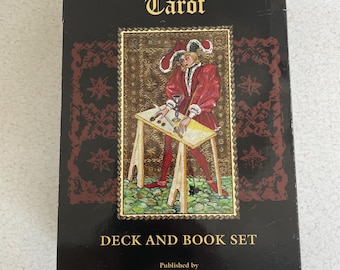 May include: A black tarot deck and book set titled "The Medieval Scapini Tarot." The box features an illustration of a person in medieval attire, and the text "DECK AND BOOK SET" is displayed prominently.