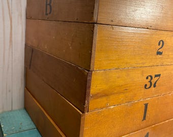 Peut inclure: Une pile de cinq boîtes de rangement rectangulaires en bois avec une finition brun clair naturel. Chaque boîte présente un identifiant noir distinct imprimé sur la face avant, incluant les caractères B, 2, 37, I et III 1, empilés verticalement.