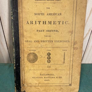 May include: An antique book titled Emerson's Second Part, The North American Arithmetic, Part Second, Uniting Oral and Written Exercises. The tan cover features black text and a diagram showing hundreds, tens, and units totaling 138.