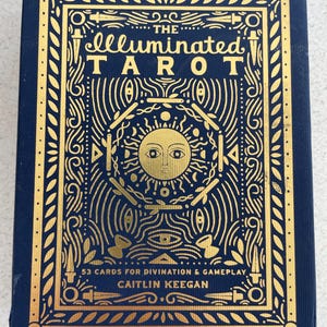 May include: A navy blue tarot card box with gold foil lettering and illustrations. The text reads "The Illuminated Tarot" with additional text indicating 53 cards for divination and gameplay by Caitlin Keegan.
