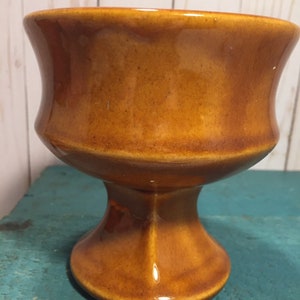 May include: A brown ceramic pedestal planter with a wide, flared top and a rounded base.