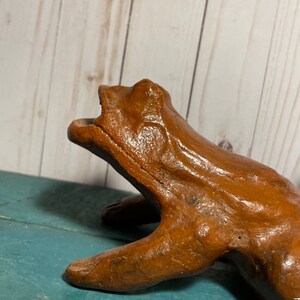May include: A brown ceramic frog figurine with a wide open mouth, sitting on a teal blue surface. The frog has a textured surface and appears to be handmade.