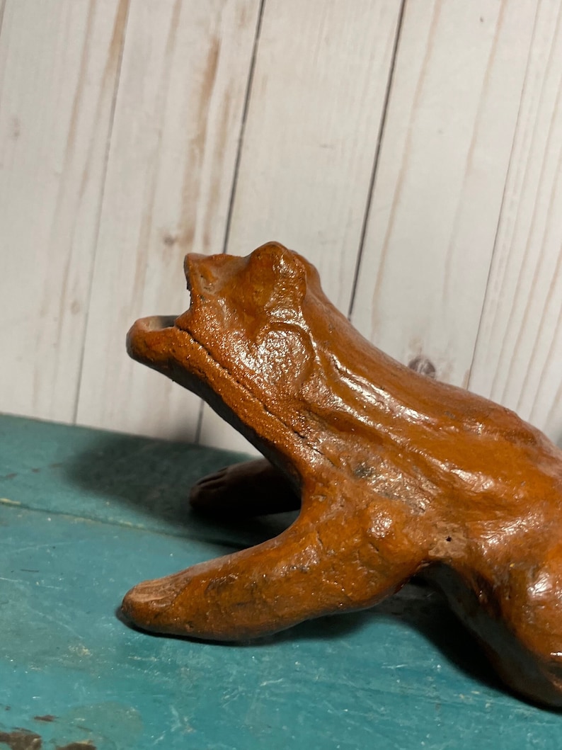 May include: A brown ceramic frog figurine with a wide open mouth, sitting on a teal blue surface. The frog has a textured surface and appears to be handmade.