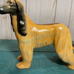 Puede incluir: Una figurita de cerámica de un perro afgano. El perro está de pie con la cabeza girada hacia un lado. La figurita está pintada en tonos marrones y negros.