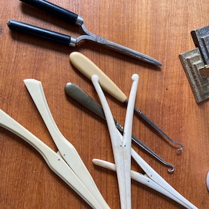 May include: A set of antique hairdressing tools, including a pair of curling tongs with black handles, a bone hair pick, and a bone hair comb. The tools are arranged on a wooden surface.