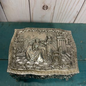 May include: A silver-plated trinket box with an ornate design featuring two women in a garden setting. The box has a hinged lid and sits on four small feet.