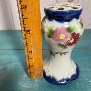 May include: A white porcelain flower frog with a blue and gold rim. The frog has a floral design with pink and red flowers. It is approximately 10 cm tall.