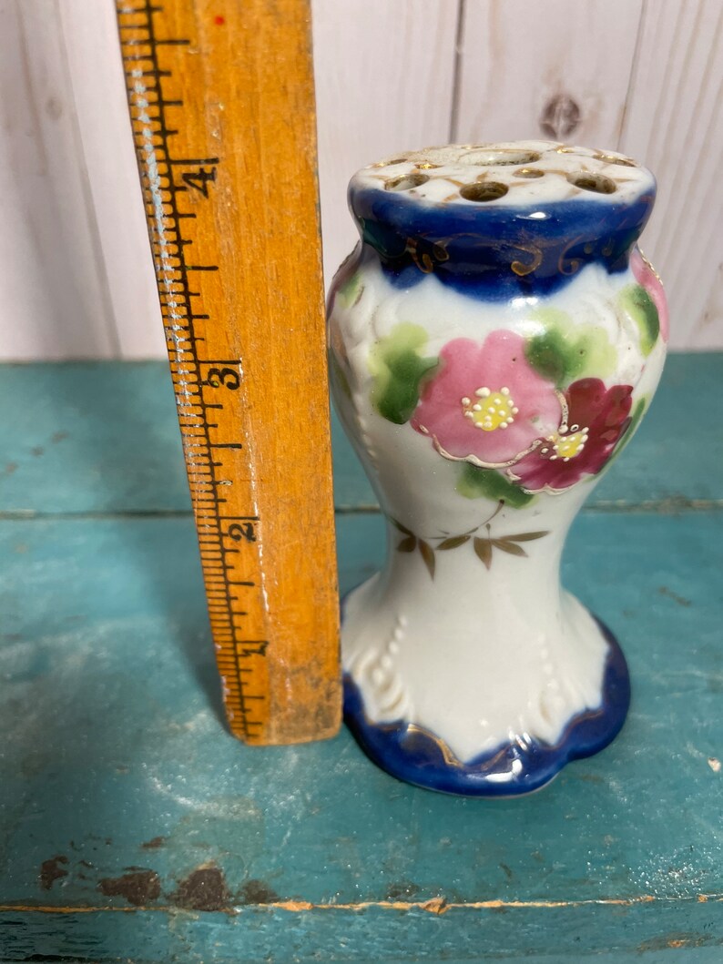 May include: A white porcelain flower frog with a blue and gold rim. The frog has a floral design with pink and red flowers. It is approximately 10 cm tall.