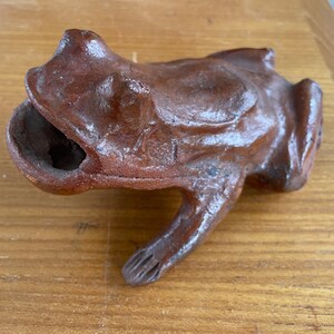 May include: A terracotta frog figurine with a textured surface and a hollowed-out mouth. The frog is brown and has a glossy finish.