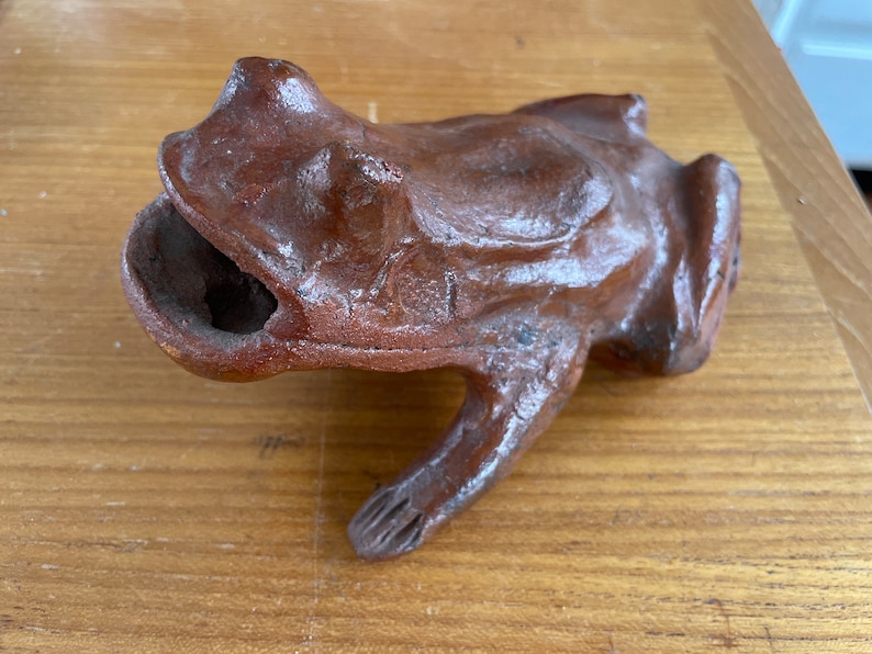 May include: A terracotta frog figurine with a textured surface and a hollowed-out mouth. The frog is brown and has a glossy finish.