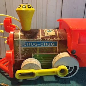 May include: Vintage wooden toy train with a red caboose, yellow wheels, and a yellow whistle. The train has a brown body with the words "CHUG-CHUG" printed on it.