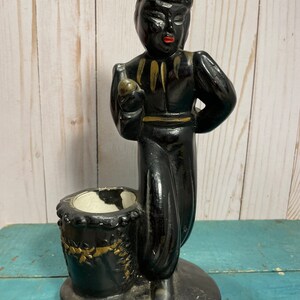 May include: Black ceramic figurine of a person holding a bottle, with a gold-trimmed hat and a gold-trimmed drum-shaped container. The figurine is standing on a heart-shaped base.