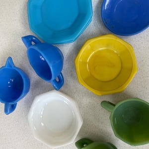 Könnte beinhalten: Eine Sammlung farbenfroher Vintage-Geschirrteile, darunter Teller und Tassen in Blau-, Gelb-, Weiß- und Grüntönen. Die Teller haben eine achteckige Form, und die Tassen haben Henkel.