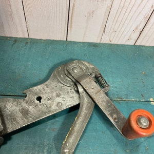 Könnte beinhalten: Ein vintage-Metall-Dosenöffner mit rotem Griff und Federmechanismus. Der Öffner hat eine silberne Oberfläche und ist so konzipiert, dass er Dosen mit einem glatten, gleichmäßigen Schnitt öffnet.