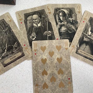 May include: A collection of playing cards featuring vintage-style illustrations. The cards display portraits of figures and scenes, with a central card showing ten golden hearts. The cards are in shades of beige, brown, and gold.