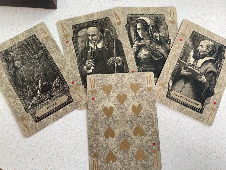 May include: A collection of playing cards featuring vintage-style illustrations. The cards display portraits of figures and scenes, with a central card showing ten golden hearts. The cards are in shades of beige, brown, and gold.