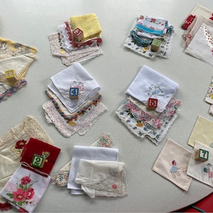 Könnte beinhalten: Eine Sammlung von Vintage-Taschentüchern mit Spitzenbesatz und gestickten Designs. Die Taschentücher sind auf einer weißen Oberfläche mit nummerierten Holzwürfeln angeordnet.