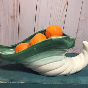 以下が含まれることがあります： 緑と白のセラミック製の豊饒の角のボウルで、カールしたハンドルが付いています。ボウルには、オレンジ色のミカンが4つ入っています。