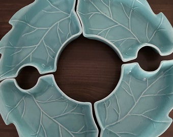 May include: Four light blue leaf-shaped ceramic dishes arranged in a circle. Each dish has a detailed vein pattern and a curved edge. The dishes are on a dark brown wooden surface.