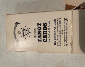 May include: A cream-colored tarot card box with black text. The box reads "TAROT CARDS" and "THE CHURCH OF LIGHT" with an address. The box also features a symbol with a star and a sun.