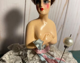 May include: A vintage pincushion doll wearing a white lace dress with blue and pink ribbons, and a pearl-topped pin. The doll has a painted face with red lips and brown hair.