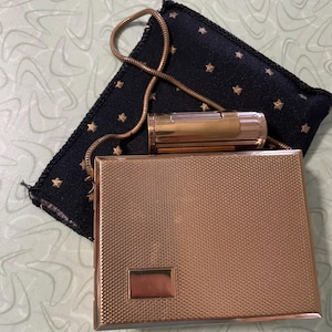 May include: A vintage gold-toned cigarette case, lighter, and a small black pouch with gold star accents. The cigarette case has a textured surface and a rectangular accent. The lighter is cylindrical and gold-toned. The pouch has a gold chain.