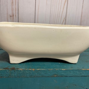 May include: A white ceramic planter with a rectangular shape and a slightly curved bottom. The planter has four short legs and is sitting on a blue wooden surface.