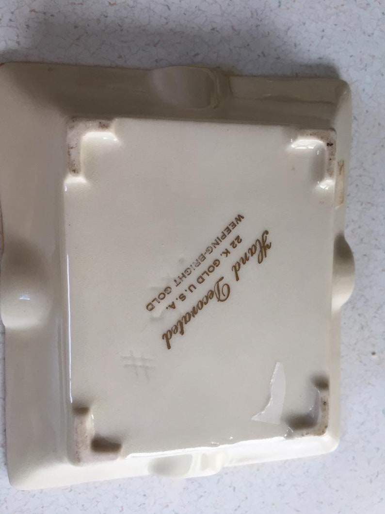 May include: A white ceramic ashtray with a gold inscription that reads "Hand Decorated 22 K Gold U.S.A.  Weeeping-Bright Gold".
