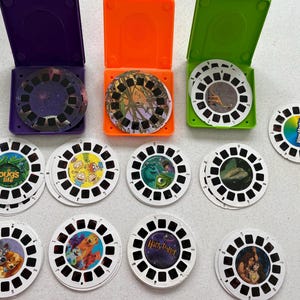 May include: A collection of View-Master reels and viewers in purple, orange, and green cases. The reels feature images from various animated movies and a color wheel reel. The cases are open, displaying the reels.
