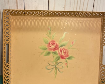 May include: A vintage rectangular serving tray with a gold filigree frame. The tray's interior has a pale peach background with a hand-painted floral design of pink roses and green leaves. A decorative bow is painted below the flowers.