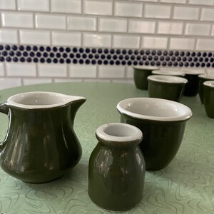 Może przedstawiać: Zestaw czterech zielonych ceramicznych kubków i dzbanka. Kubki są różnych rozmiarów i mają białe obramowanie. Dzbanek ma białe obramowanie i uchwyt. Kubki i dzbanek stoją na zielono-białej powierzchni.