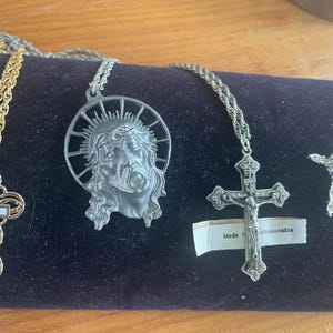 Könnte beinhalten: Vier silberfarbene religiöse Halsketten mit verschiedenen Anhängern. Ein Anhänger ist ein Kreuz mit einer Figur von Jesus Christus, ein anderer ist ein Kreuz mit einem Blumendesign, ein weiterer ist ein Kreuz mit einem einzelnen Stein und der letzte ist ein Anhänger mit einer Darstellung des Gesichts von Jesus Christus.