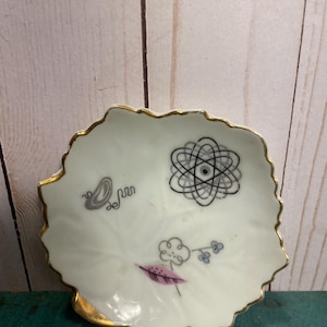 Könnte beinhalten: Eine weiße Porzellanschale mit goldfarbenem Rand und einem blattförmigen Design. Die Schale zeigt ein schwarz-weißes Atom-Symbol, eine rosa Feder und einen grauen Schmetterling.