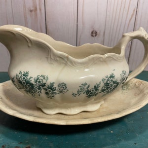 May include: A vintage white gravy boat with a green floral design and a matching saucer. The boat has a scalloped edge and a handle.
